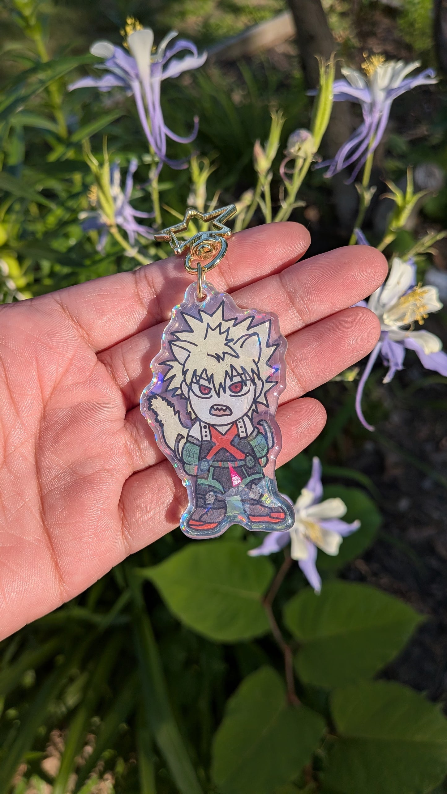 Hand holding a colorful keychain of a chibi Katsuki Bakugo with cat ears in front of green plants and flowers.