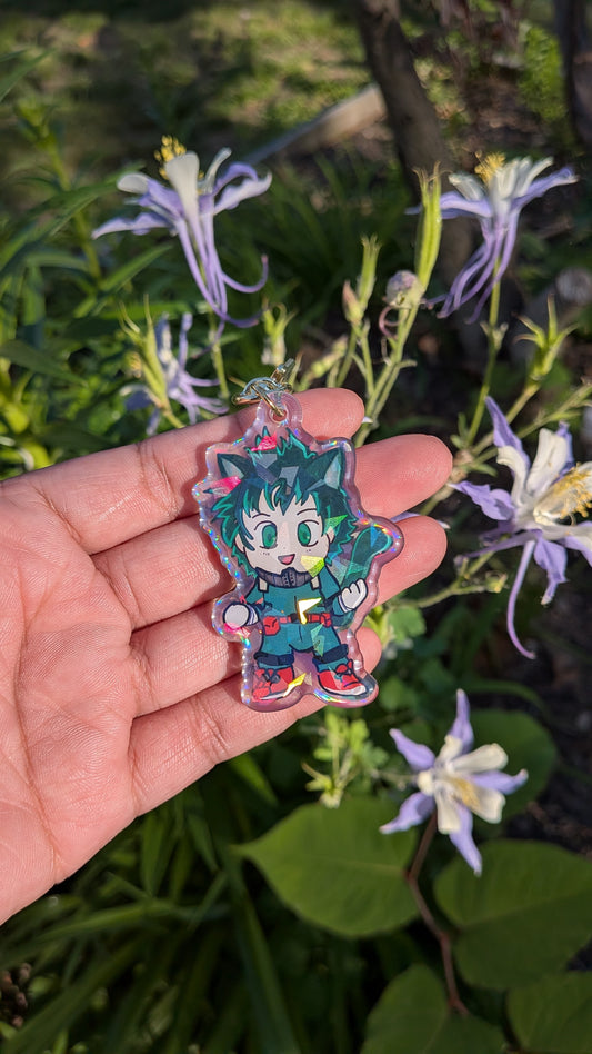 Hand holding a colorful character charm of a chibi Izuku Midoriya with cat ears  in front of green foliage and flowers
