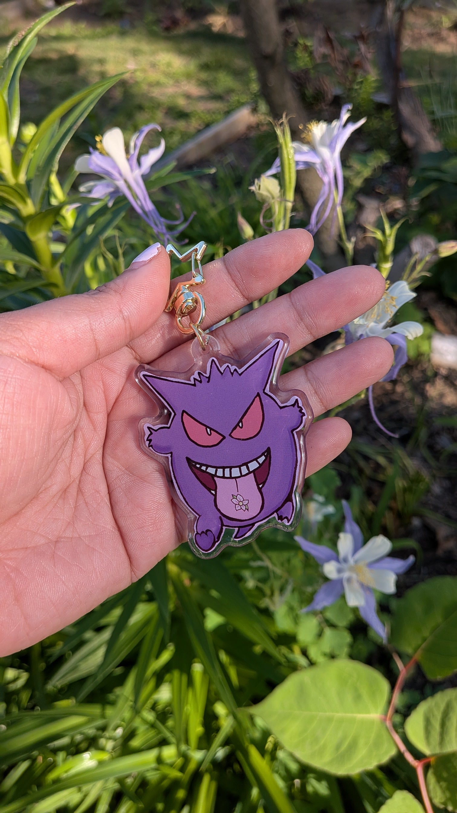 Hand holding a purple cartoon character keychain with flowers and greenery in the background