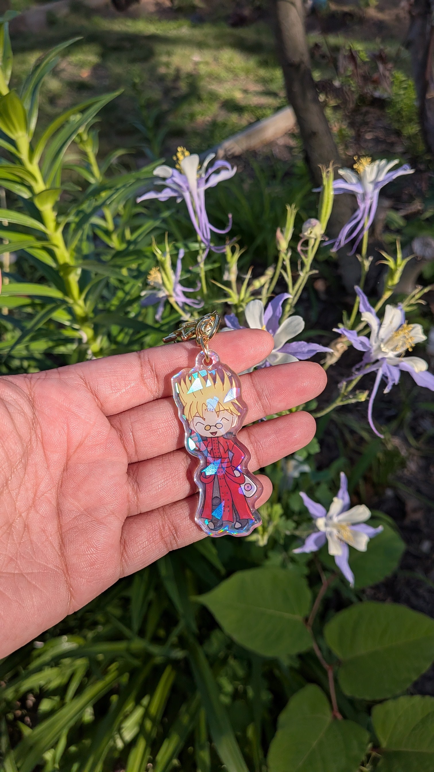 Hand holding a colorful keychain with a chibi design of vash stampede  in front of green foliage and flowers.