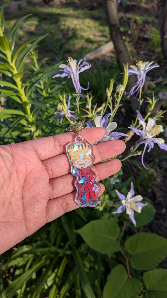 Hand holding a colorful keychain with a chibi design of vash stampede  in front of green foliage and flowers.