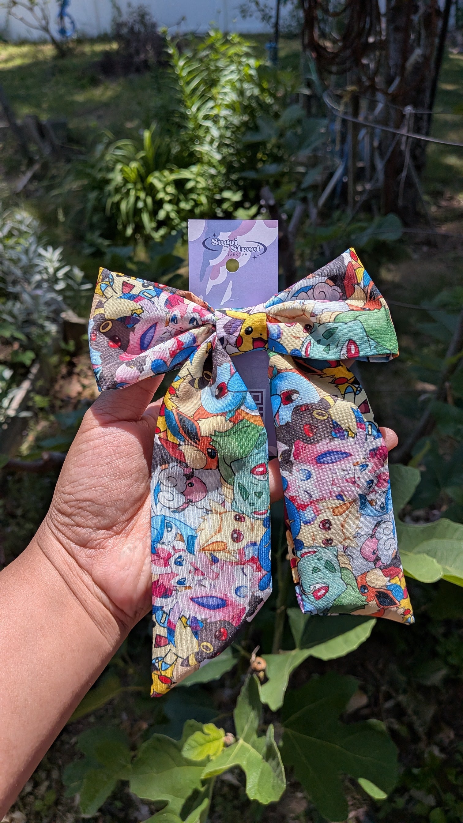 Colorful fabric bow with a pokemon pattern held in a hand with a blurred garden background